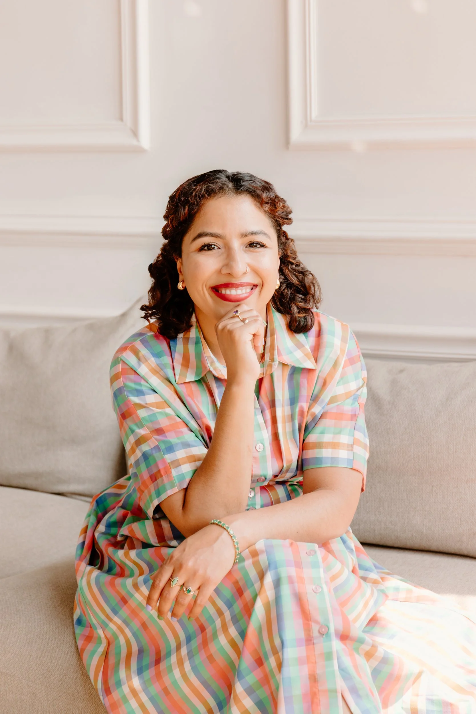 EMDR and DBR therapist Jennifer Budhan seated in a bright, welcoming office, smiling warmly with a hand resting on her chin.