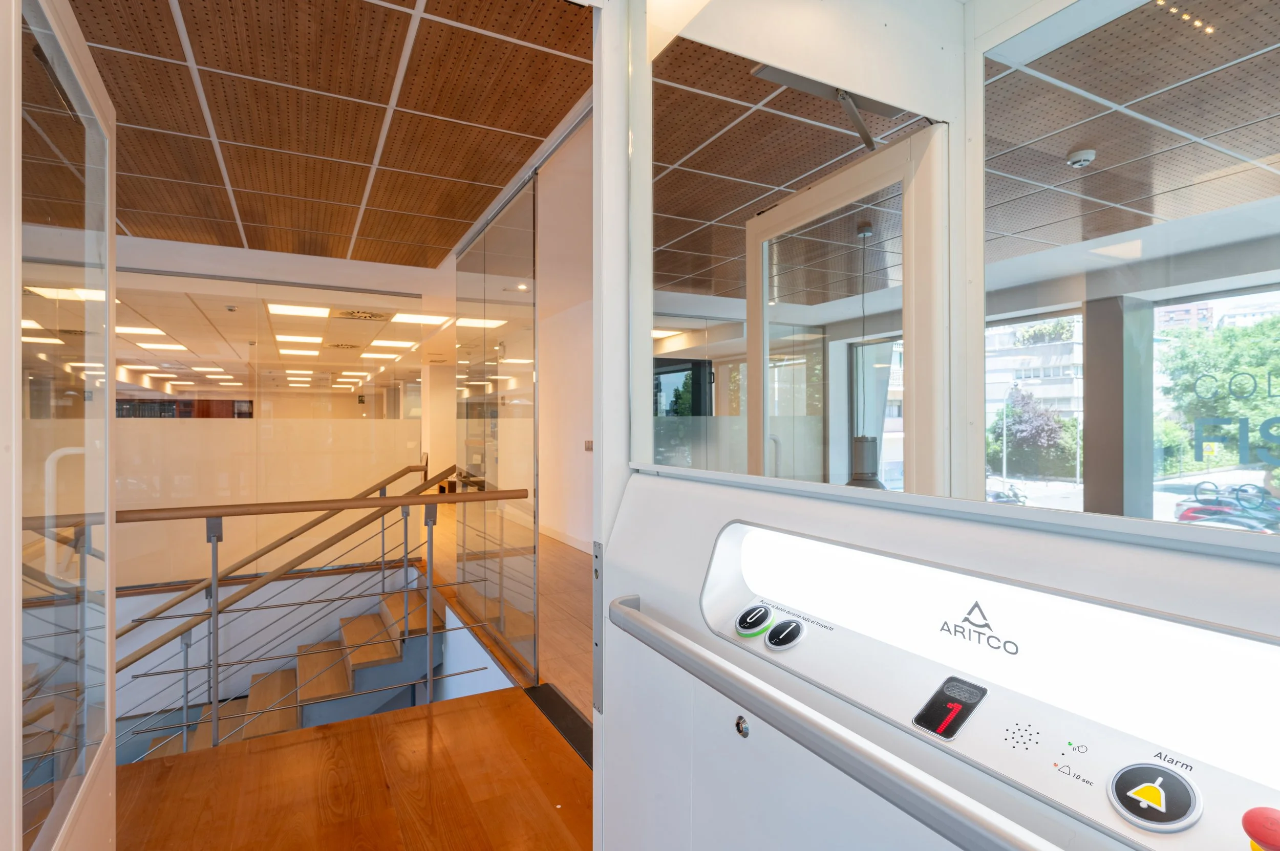 Inside a building with glass walls and wooden ceilings, an elevator control panel with buttons and an alarm indicator, near a staircase with metal railings and wooden steps, and a view of trees and buildings through the windows.