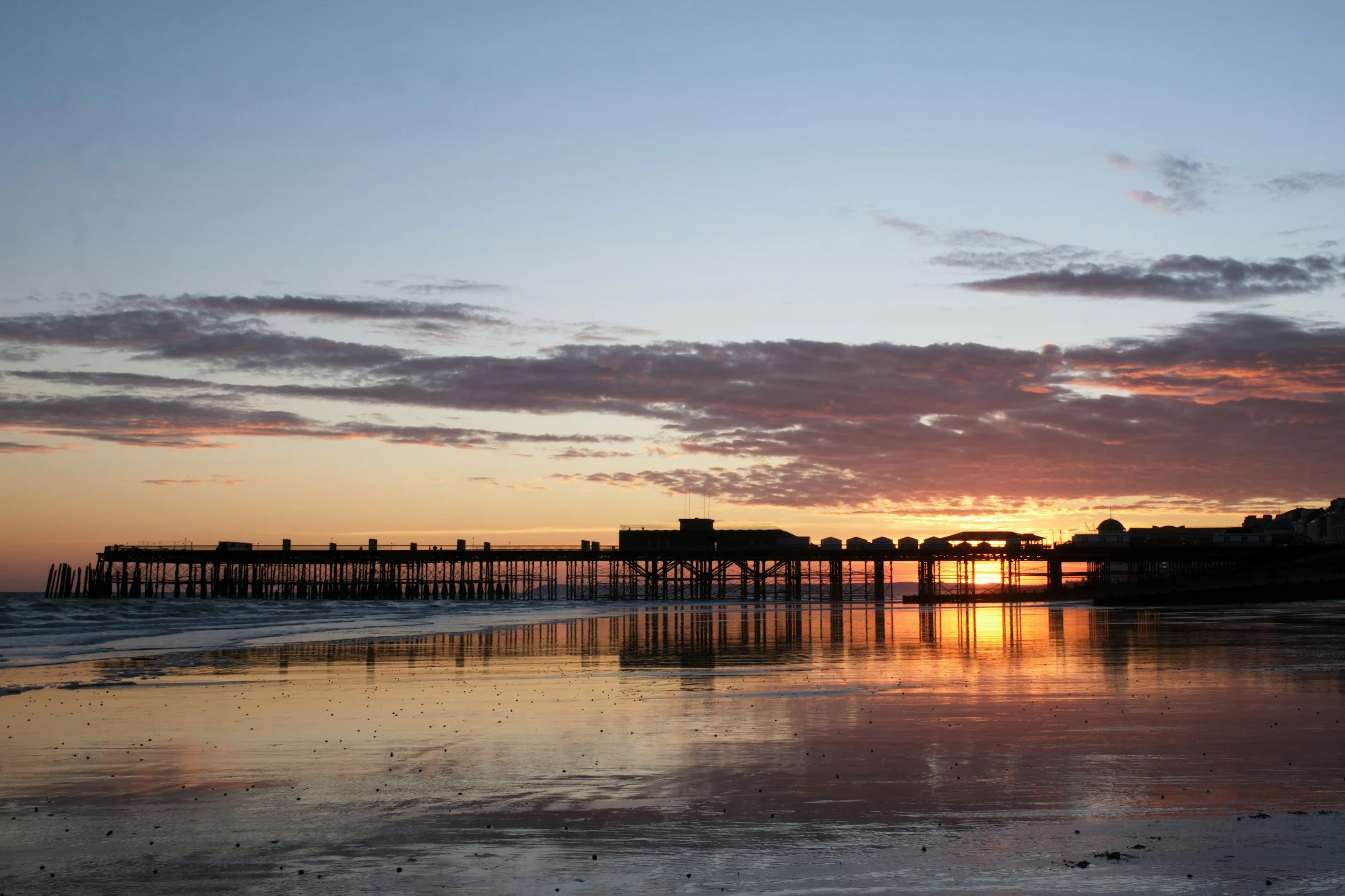 Sunset over a pier at the beach, with reflections on wet sand, partly cloudy sky with orange and pink hues.