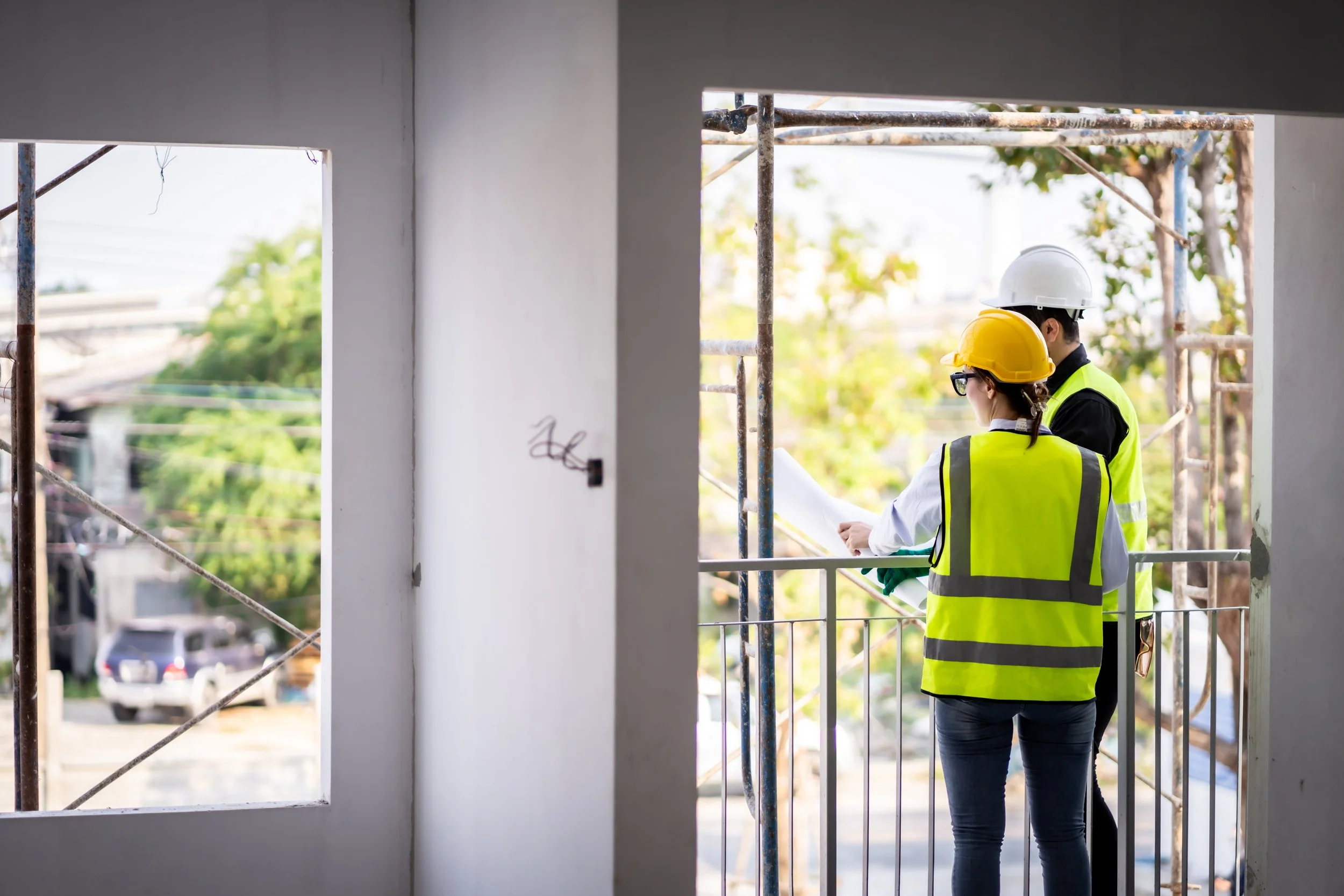 Two construction workers wearing safety helmets and reflective vests examining blueprints on a balcony at a construction site.