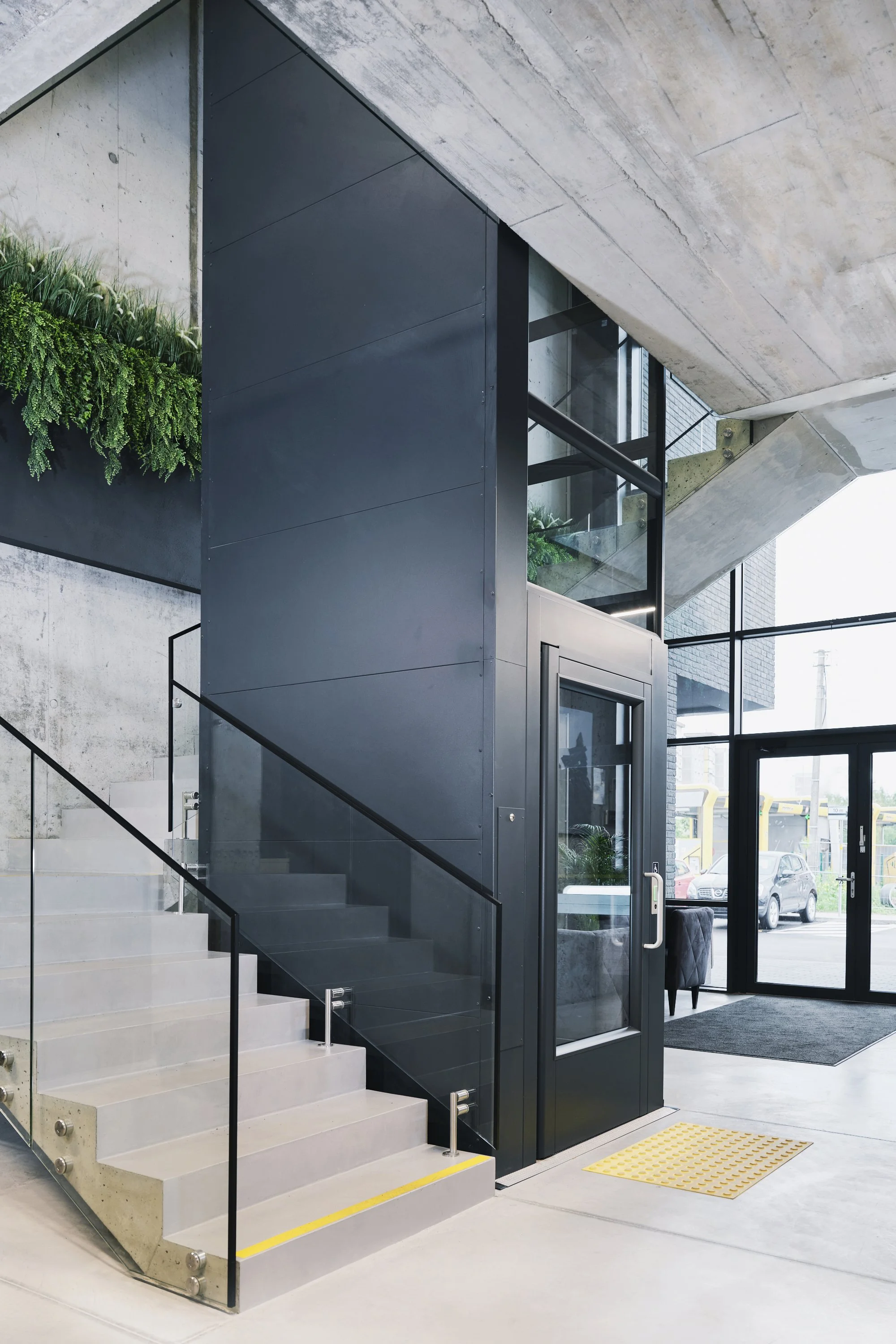 Modern building entrance with a glass door, concrete stairs with glass railing, and interior greenery.