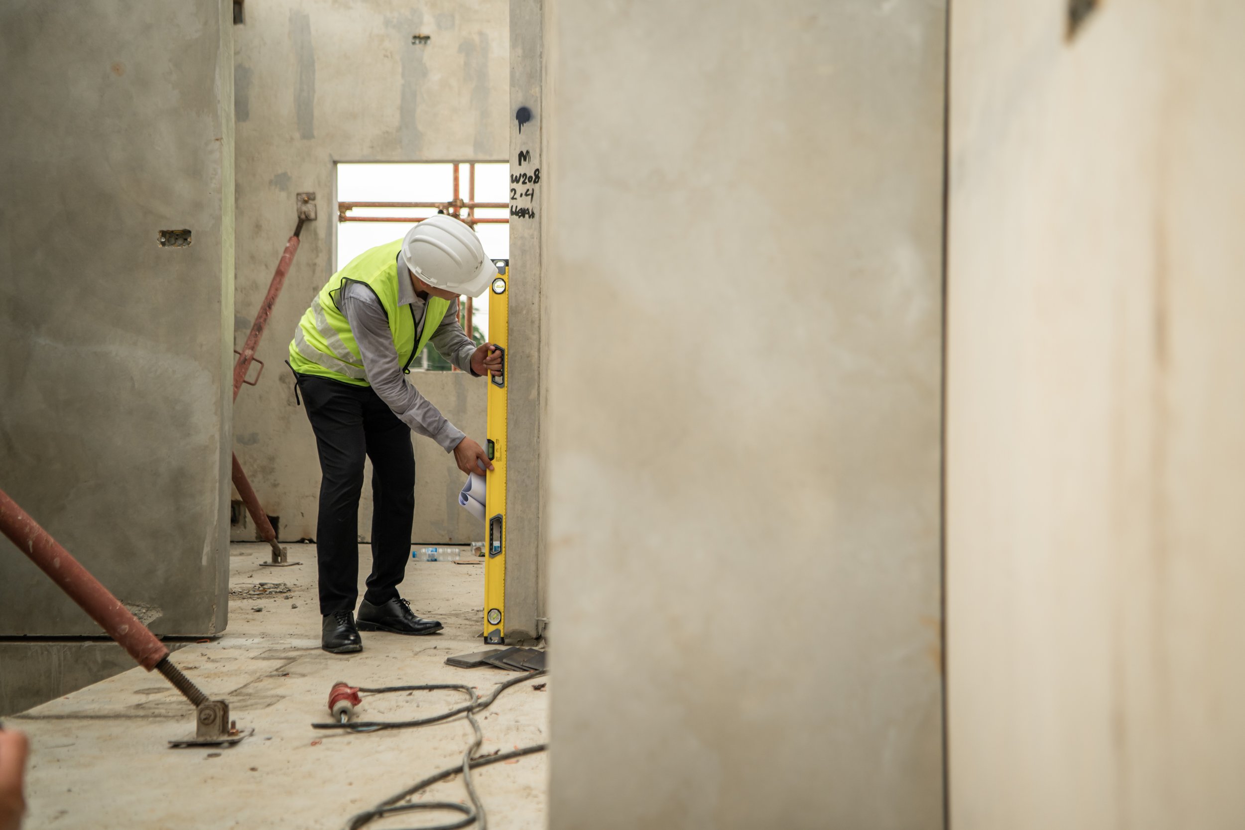 A construction worker wearing a hard hat and safety vest using a level to check the wall inside a building under construction.
