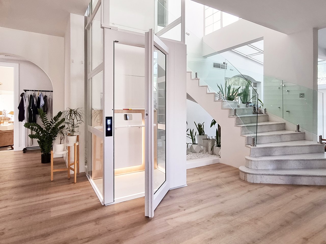Modern interior with a glass elevator, wooden stairs with glass railing, and indoor plants, featuring a light wood floor and white walls.