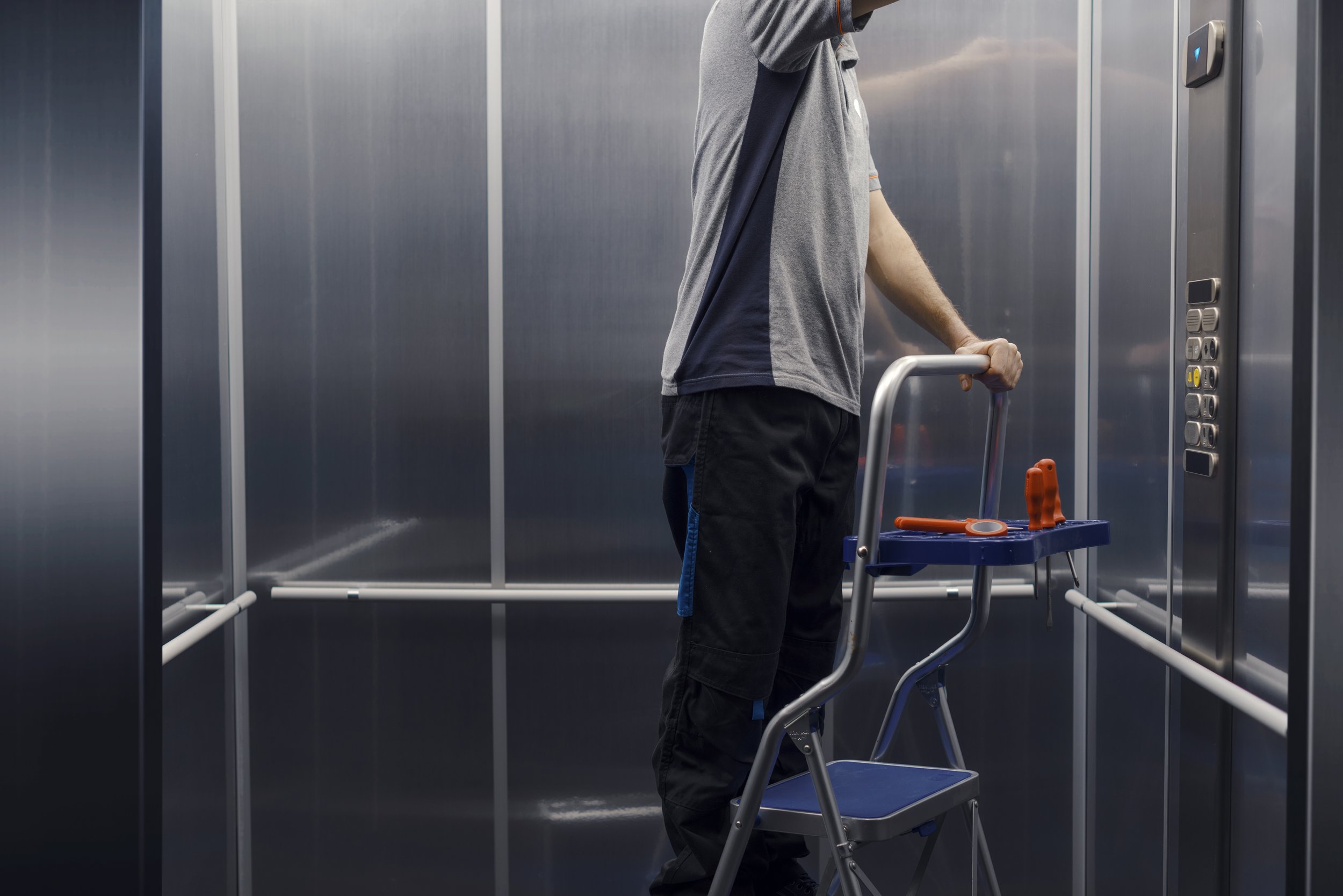 Man standing in an elevator with a step ladder and tools, facing the control panel.