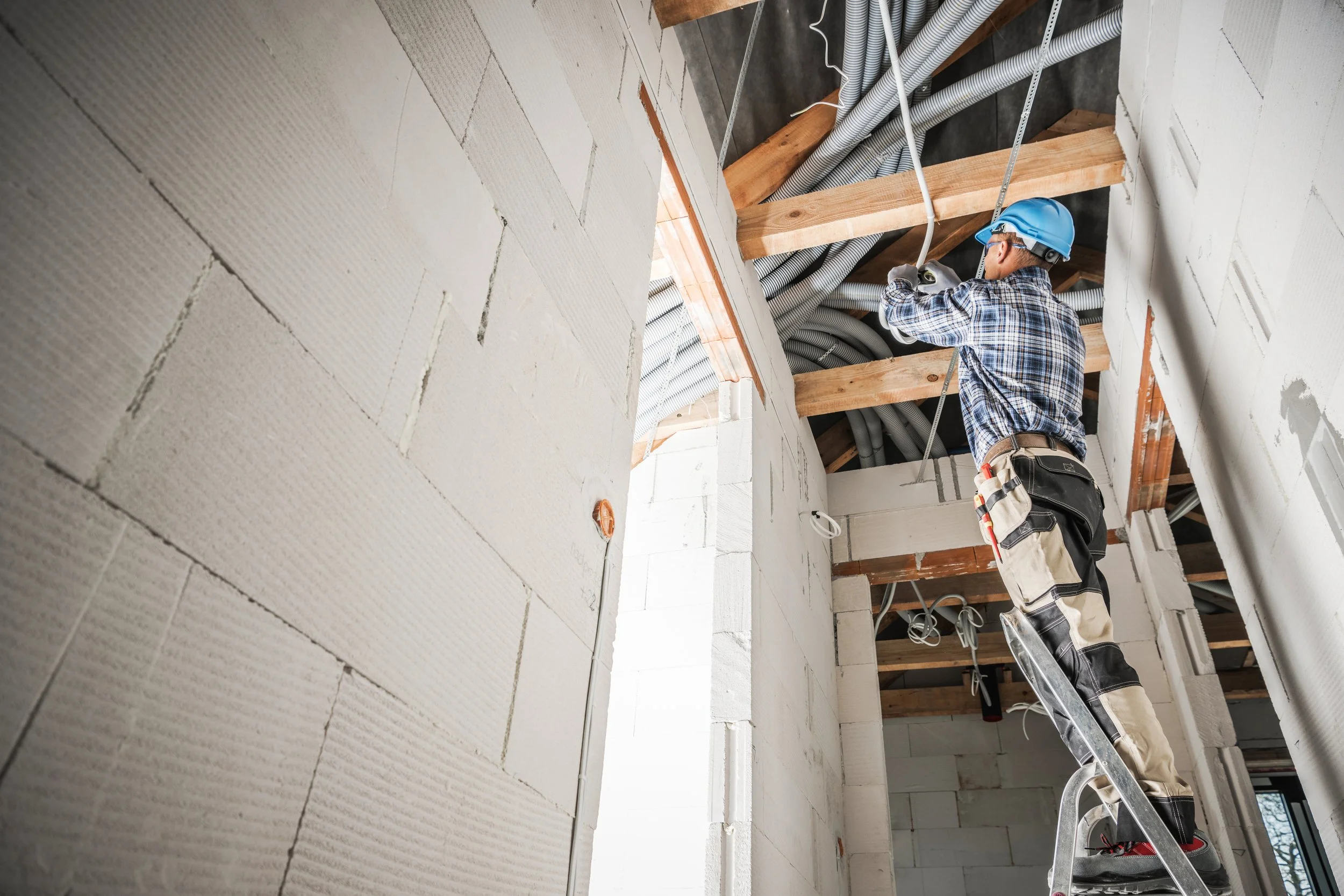 Construction worker wearing a blue helmet and safety gear on a ladder installing electrical wiring in a building with exposed ceiling pipes and wooden support beams.