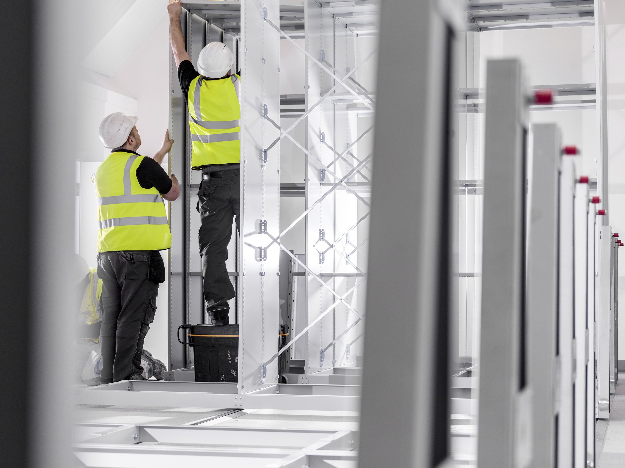 Two construction workers in safety vests and helmets installing or inspecting equipment in an industrial or construction setting with metal framing.