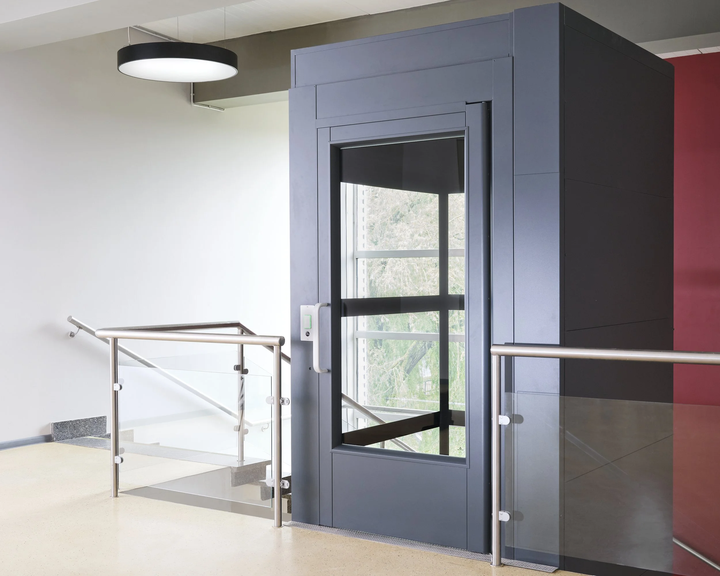 Modern glass elevator with gray exterior and glass doors, located inside a building near a staircase with stainless steel and glass railings, next to a white wall and a red wall, with a round black ceiling light.