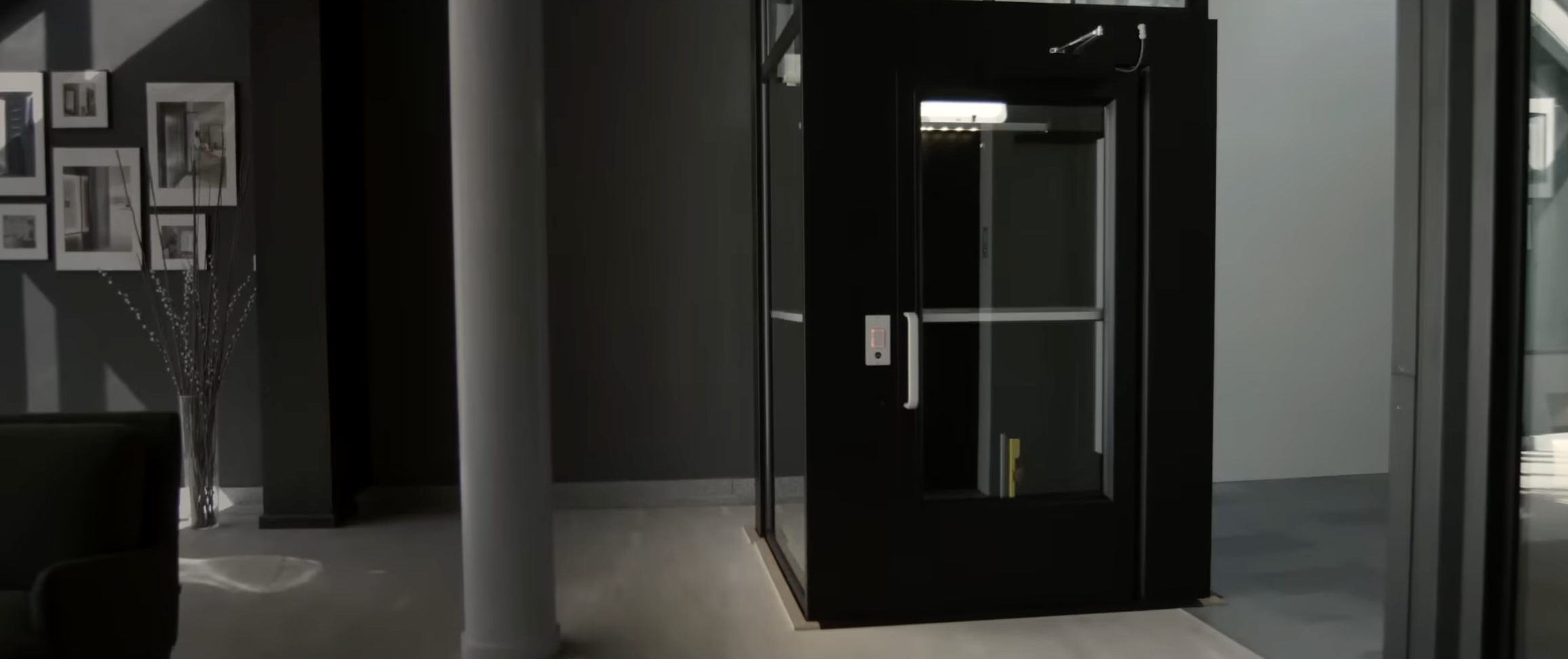 Modern glass elevator in a contemporary office or lobby with gray walls and framed photographs on the wall.