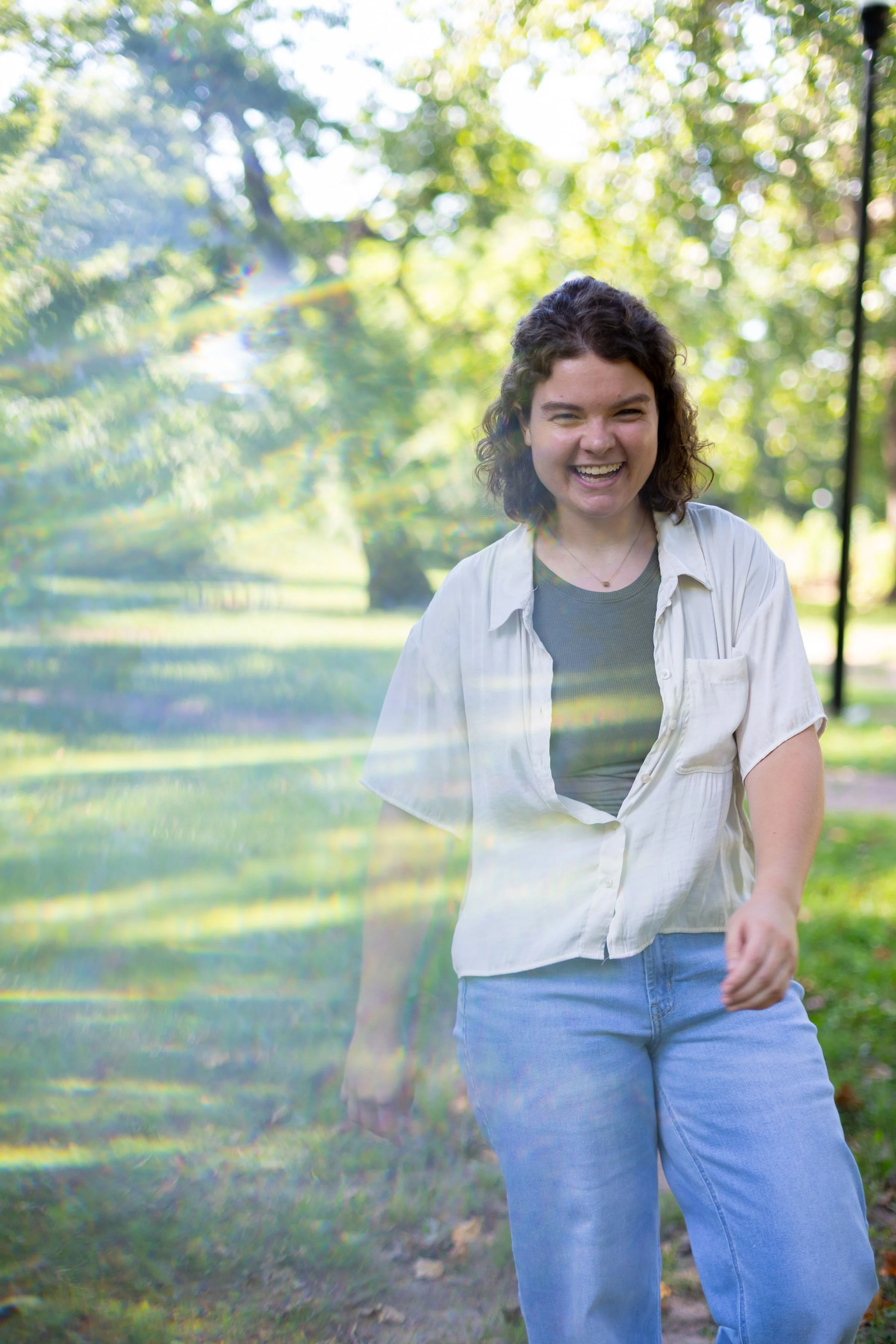 A young woman with curly hair smiling and walking outdoors in a park with green trees, wearing a white shirt and blue jeans.