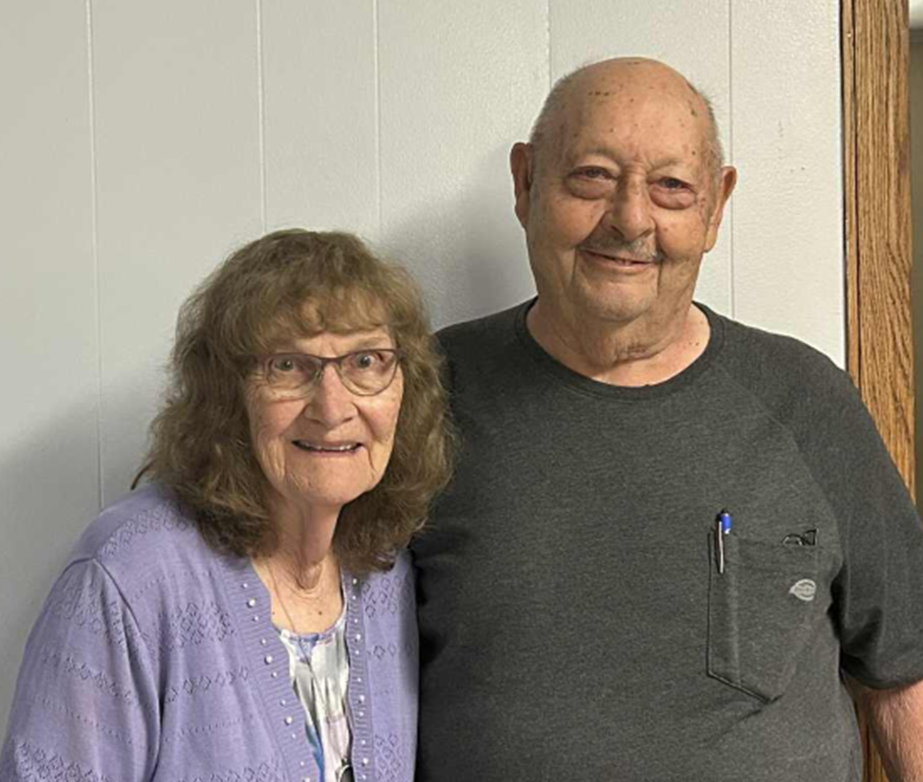 A smiling elderly woman with glasses and curly hair standing next to an elderly man with a bald head. They are indoors against a plain wall, and both are facing the camera.