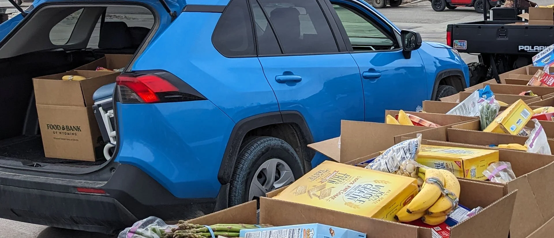 A blue vehicle parked at a food bank, with its trunk and nearby table filled with boxes of groceries including bananas, pasta, and other food items.