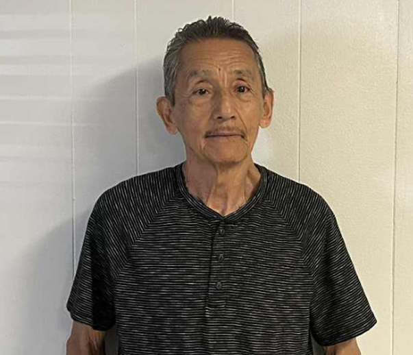 An elderly man with short gray hair standing against a plain wall, wearing a black and gray striped short-sleeve shirt.