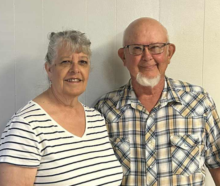 An elderly couple standing side by side, smiling at the camera, against a plain wall. The woman has short gray hair, wears a white and navy striped shirt, and earrings. The man is bald with a white beard, wears glasses, and has on a plaid button-up shirt.