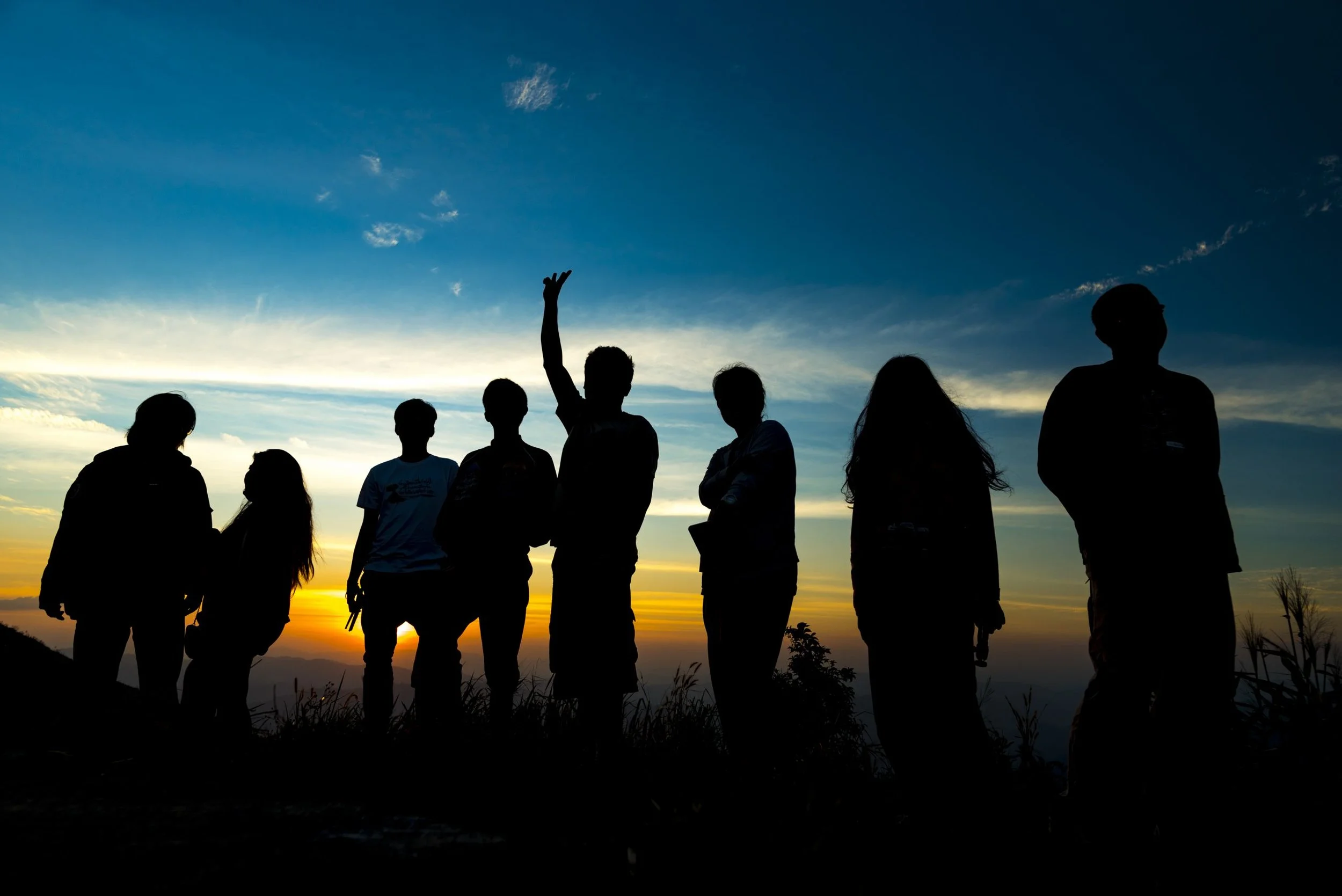 Group of seven people standing outdoors during sunset, silhouetted against a colorful sky with clouds.