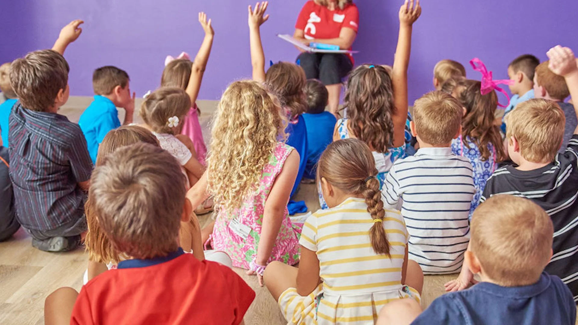 A classroom with young children sitting on the floor, many raising their hands, facing a woman in red shirt and black pants holding a clipboard, against a purple wall.