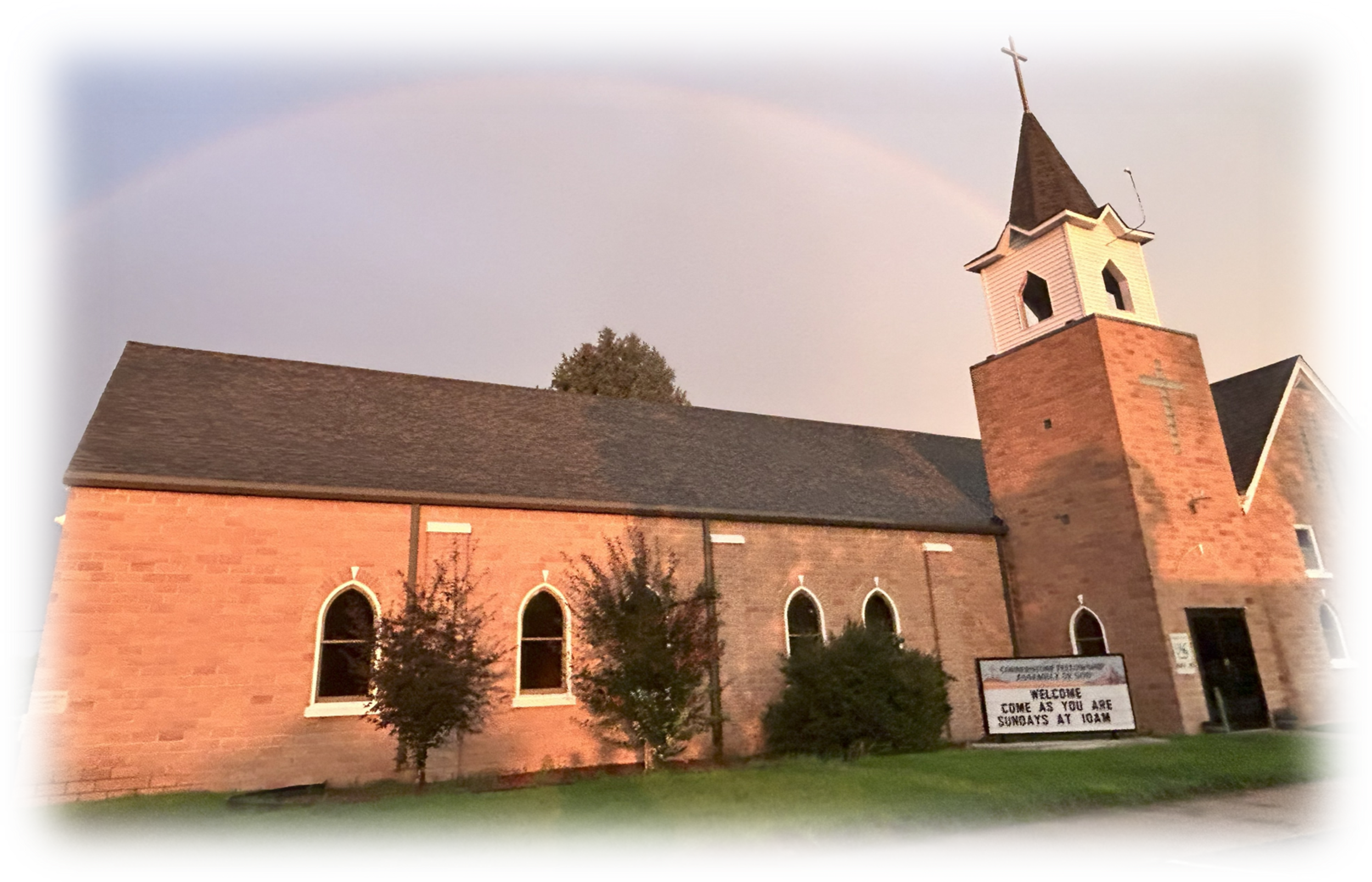A brick church with a steeple and cross, multiple arched windows, a sign welcoming visitors on a green lawn with a rainbow in the sky above.