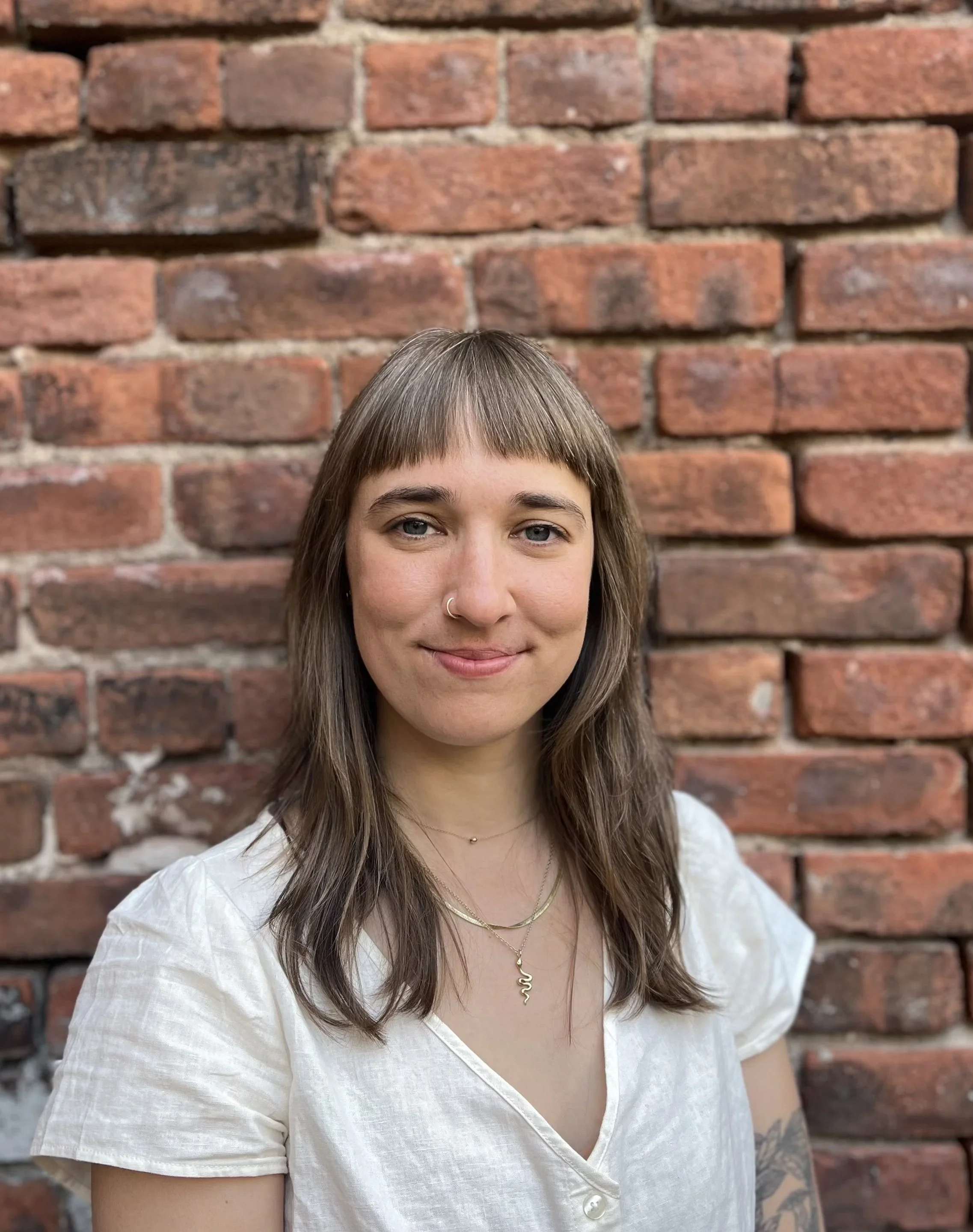 A woman with light brown hair and bangs standing in front of a brick wall, wearing a white short-sleeved shirt and jewelry.