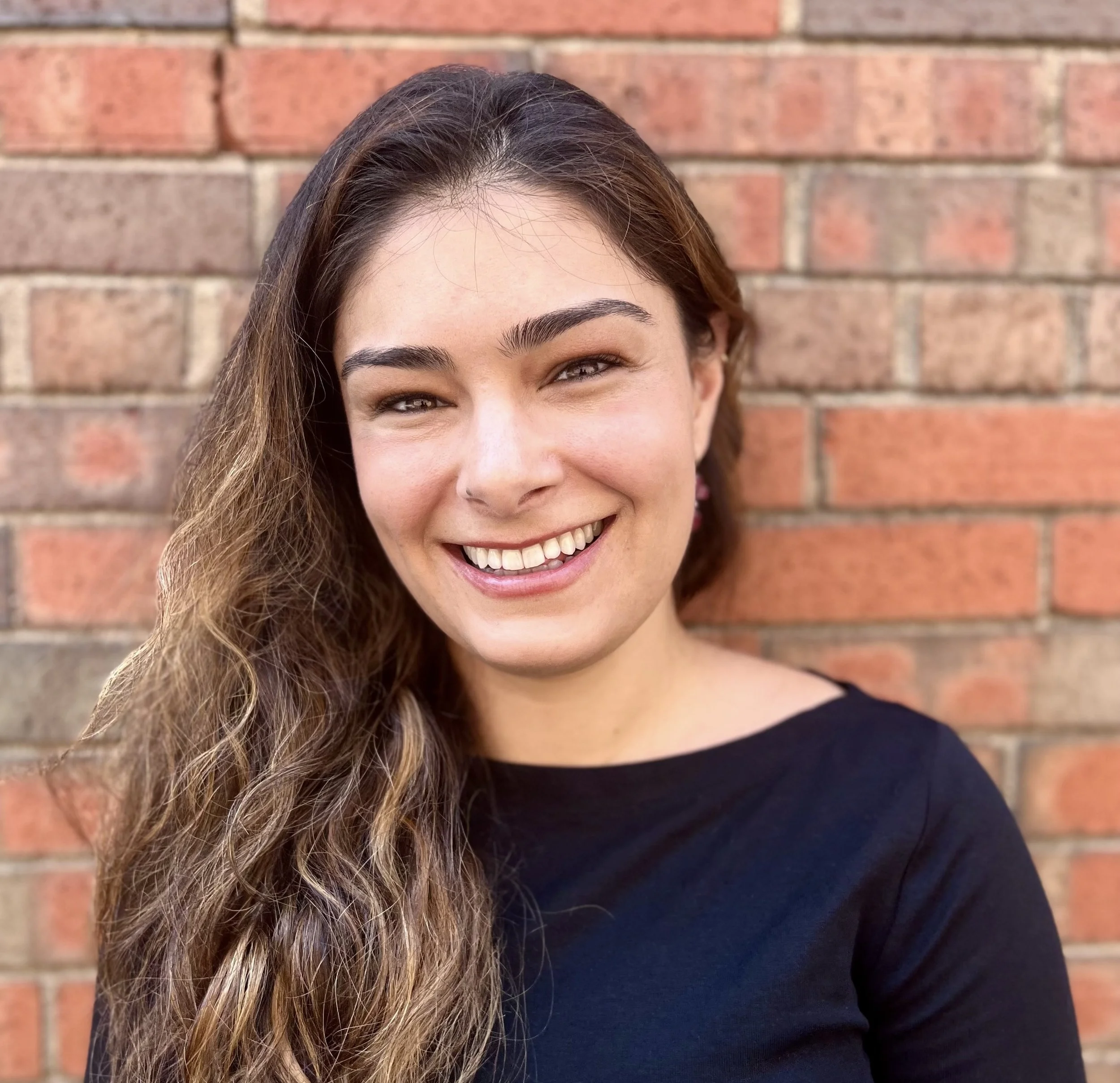 Close-up of a smiling woman with long brown hair and eyebrows, wearing a black top, standing against a red brick wall.