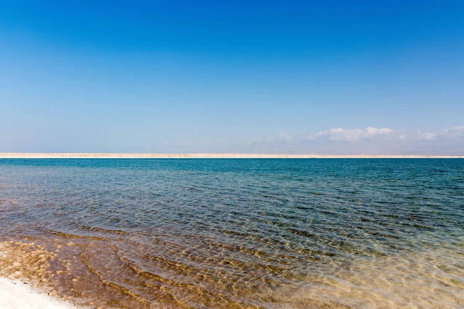 Clear turquoise water over a pale salt shore with hazy mountains on the horizon