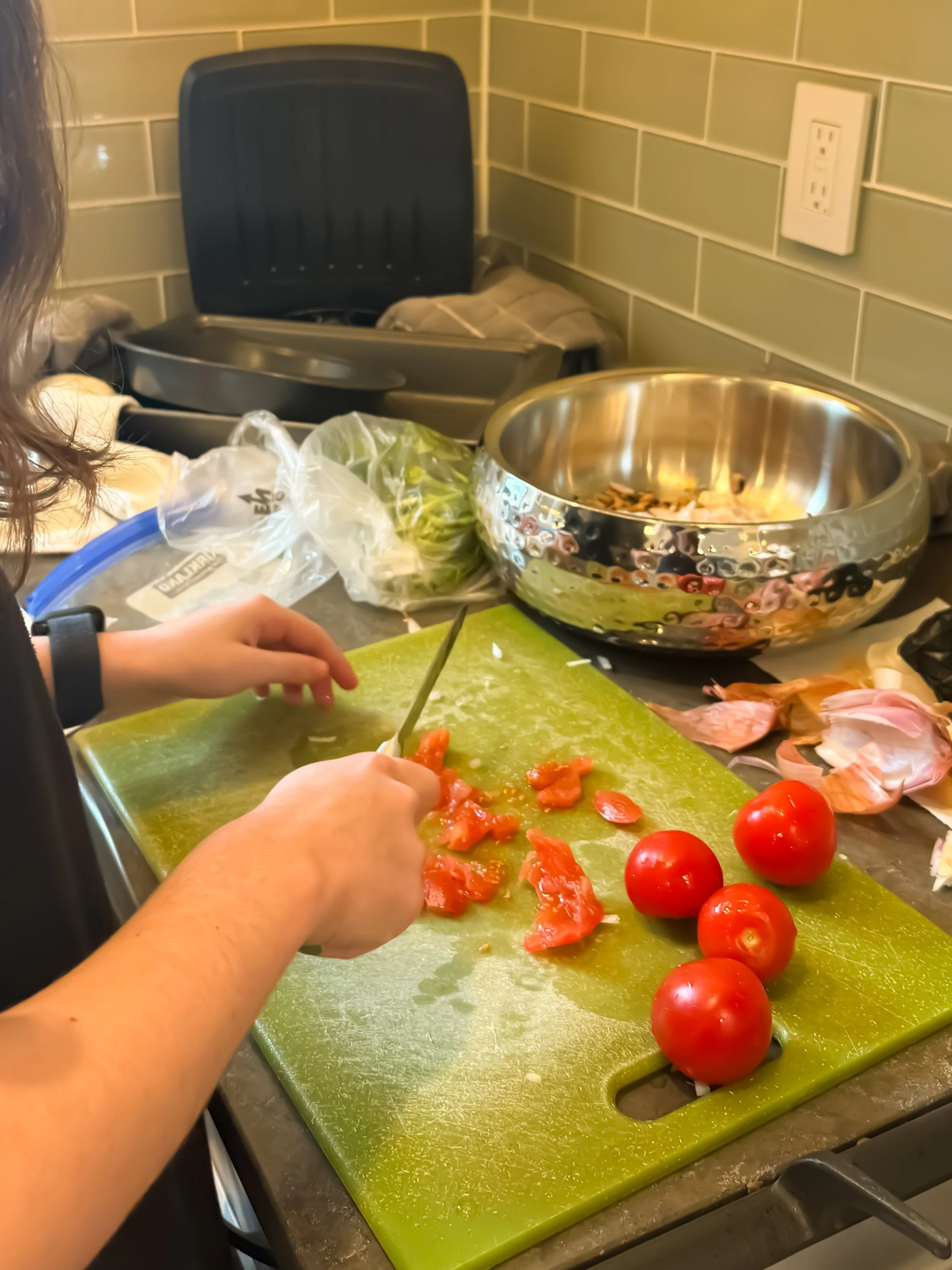Person chopping tomatoes on a cutting board in a kitchen, with whole and chopped tomatoes and cooking items on the counter.