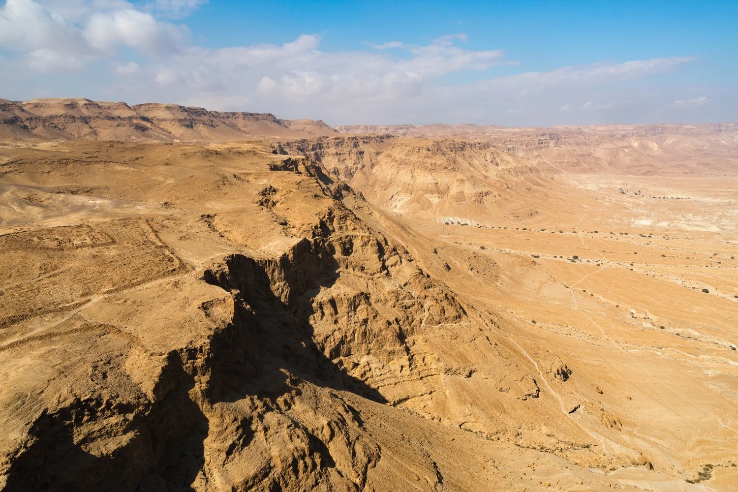 View from the top of Masada over a deep desert ravine in low golden light with a long shadow and drifting clouds