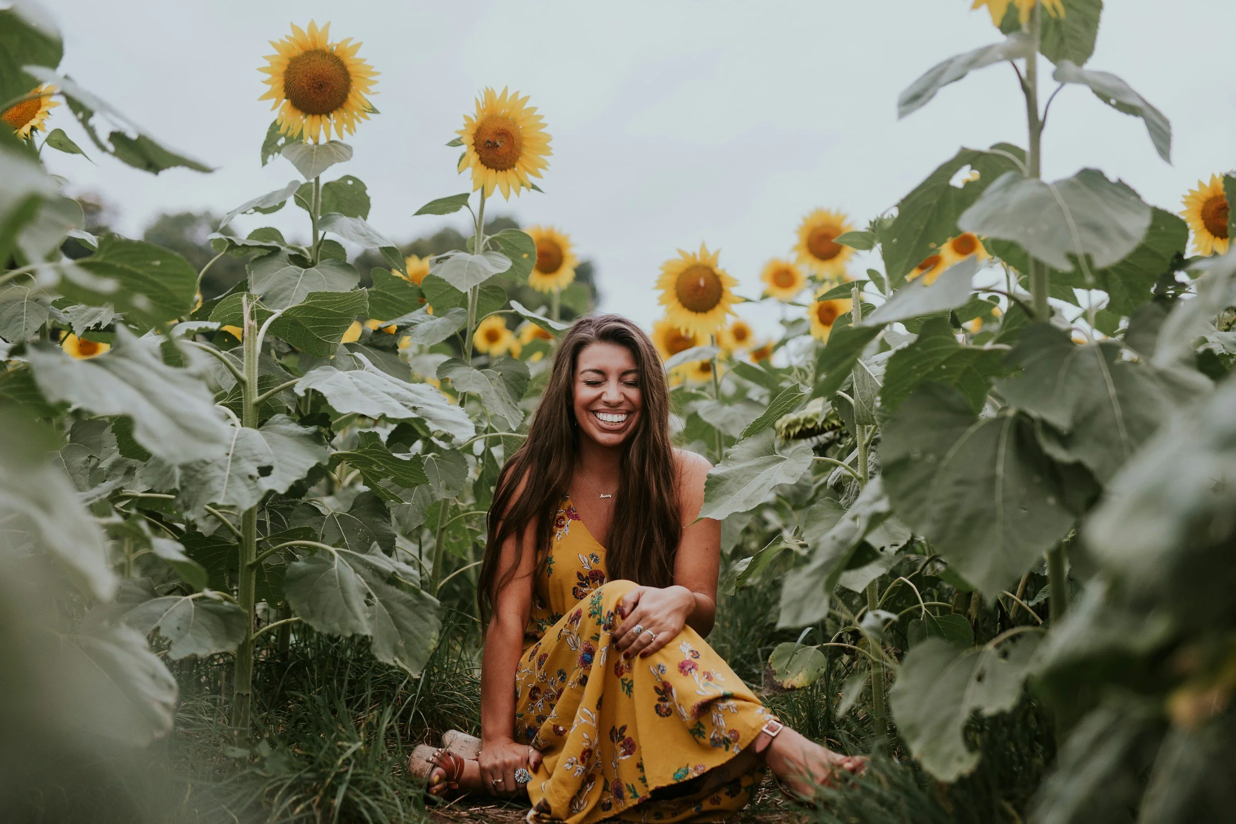 A woman in a yellow floral dress sitting among tall sunflowers, smiling and laughing.