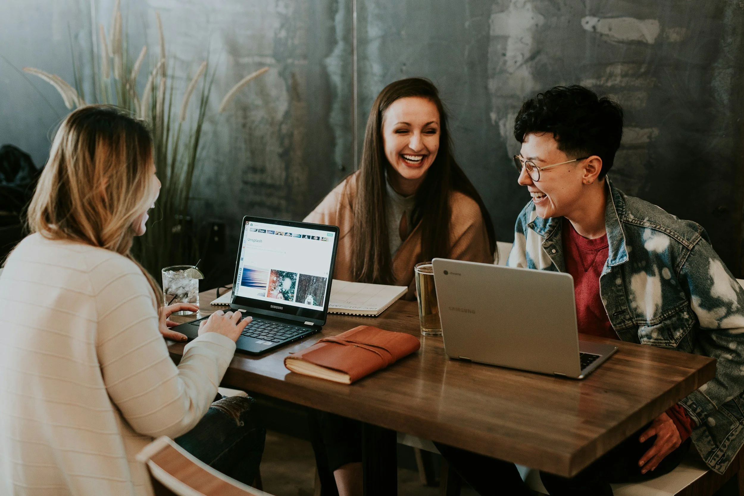 Four people sitting at a wooden table with laptops, notebooks, and drinks, laughing and enjoying a conversation in a casual setting with a gray wall and tall plant in the background.