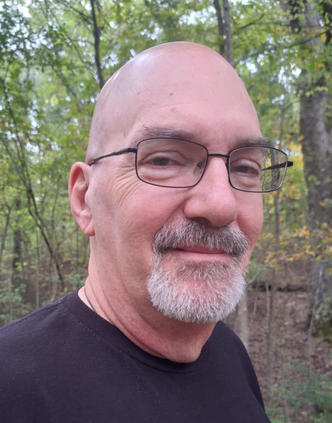 Neal Cox with glasses and a beard smiling outdoors in a forest with green trees.
