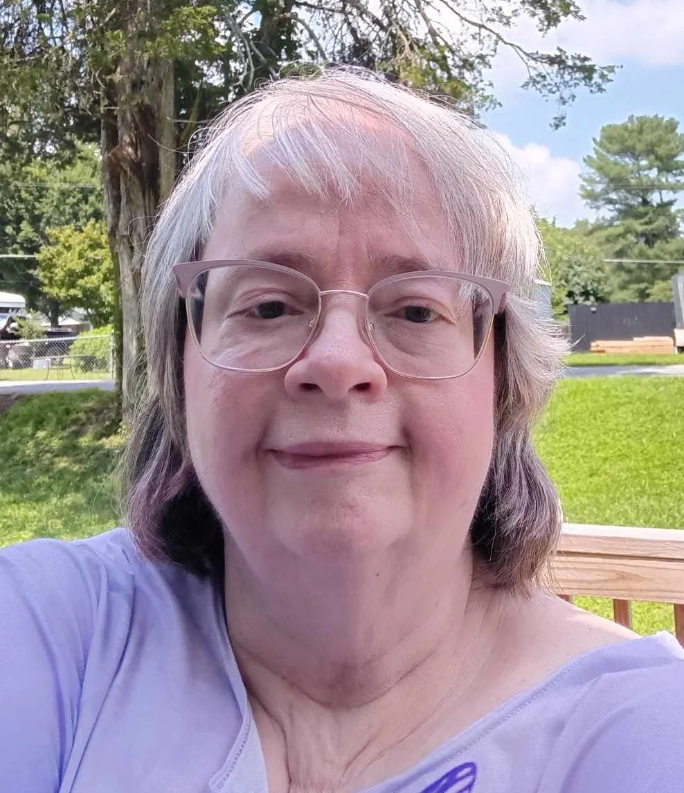 Coach Jill Salmen with glasses and shoulder-length hair taking a selfie outdoors on a sunny day, with trees and a grassy area in the background.