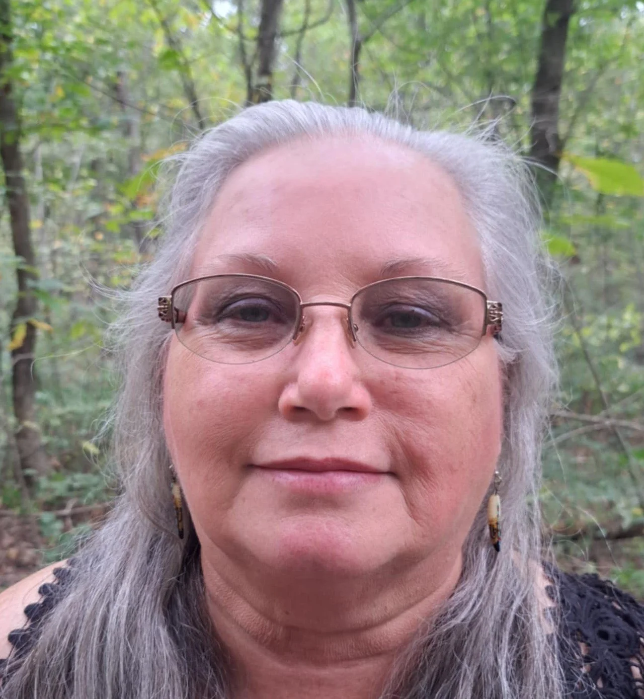 Jennifer Cox Close-up of a woman with gray hair, glasses, and earrings, standing outdoors in a wooded area.