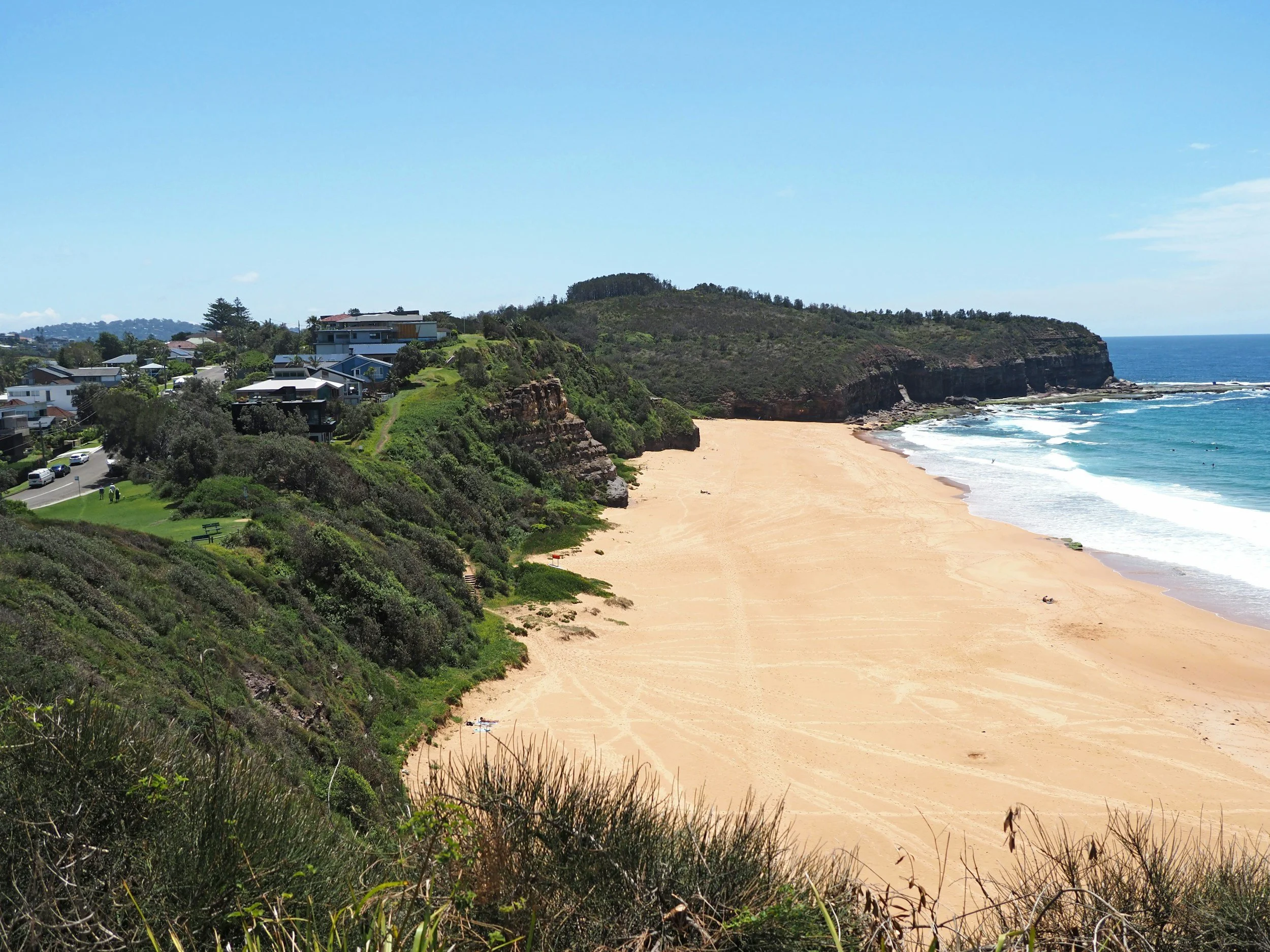 A scenic view of a beach with sandy shore, ocean waves, and cliffs, with houses on a hillside and a clear blue sky.