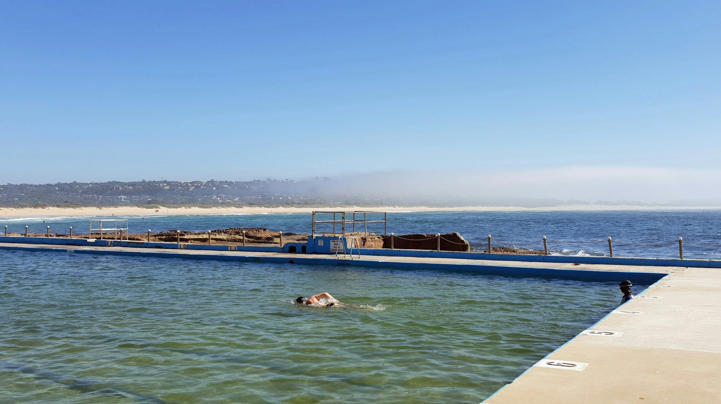 A person swimming in a pool near the ocean with a beach and houses in the background on a clear, sunny day.