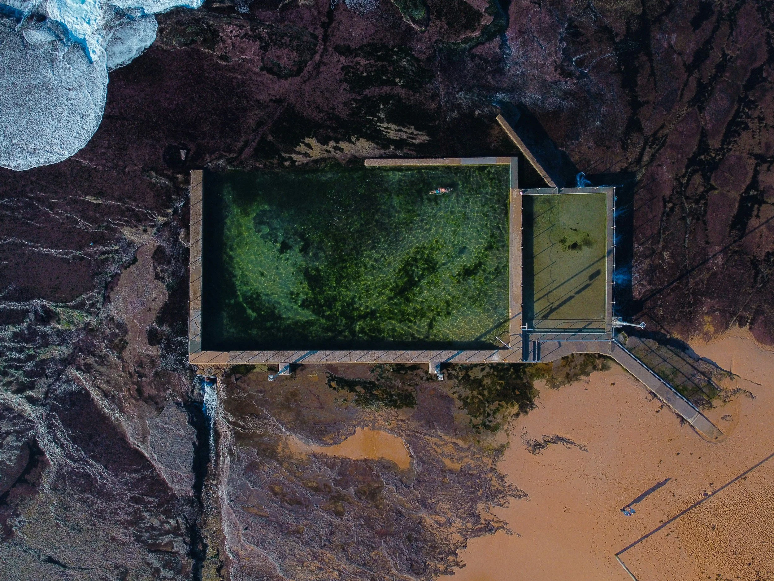 An aerial view of a waterfall flowing into a basin surrounded by rugged landscape, with a person near the edge and some structures nearby.