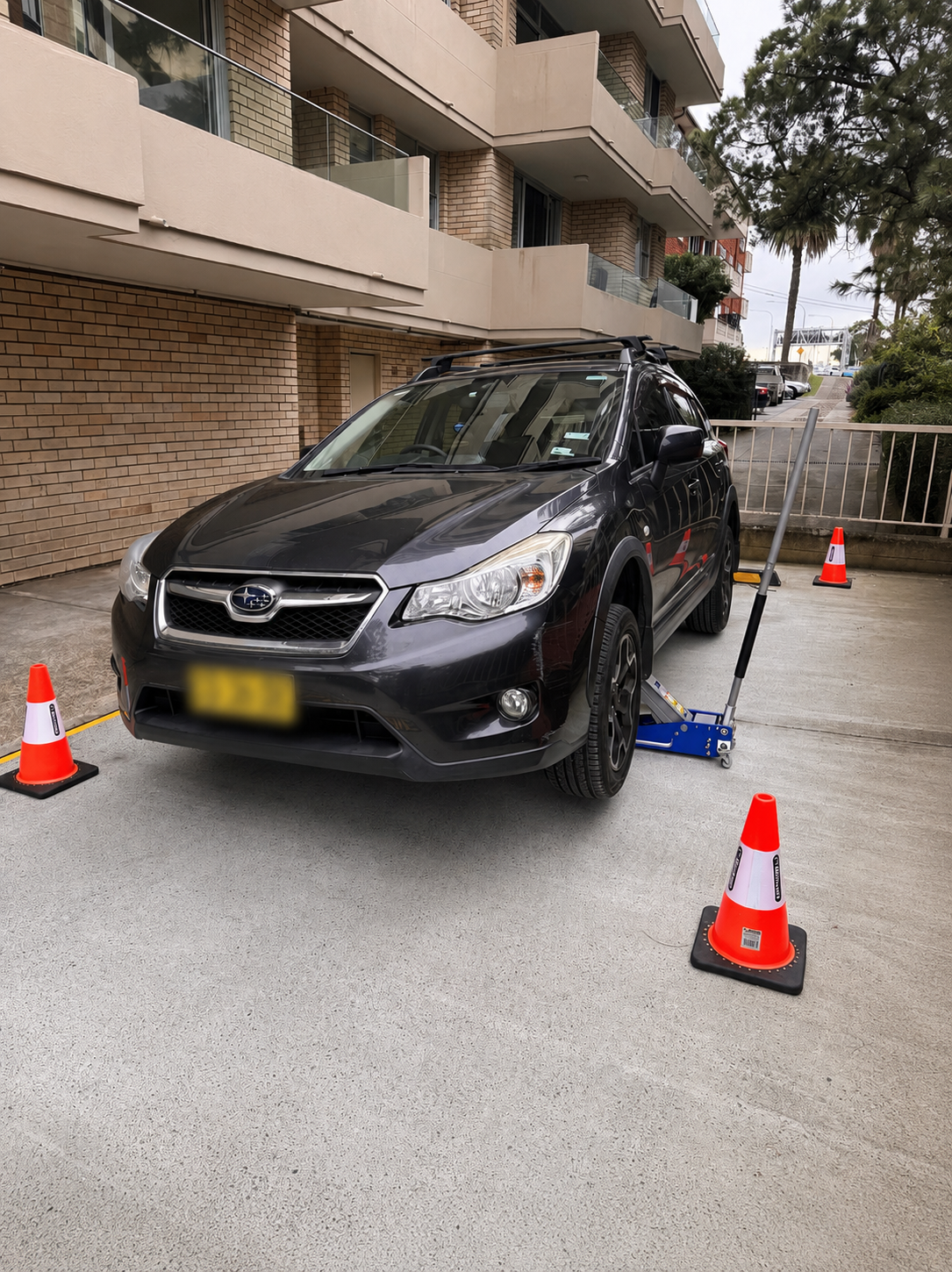 Black Subaru car parked with a wheel alignment or repair lift underneath. Four orange cones surround the vehicle, marking the work area.