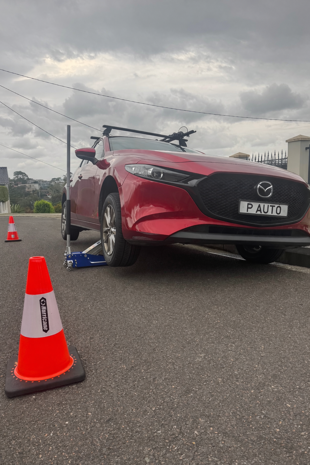 Red Mazda car on a wheel alignment lift with orange cones nearby, under a cloudy sky.