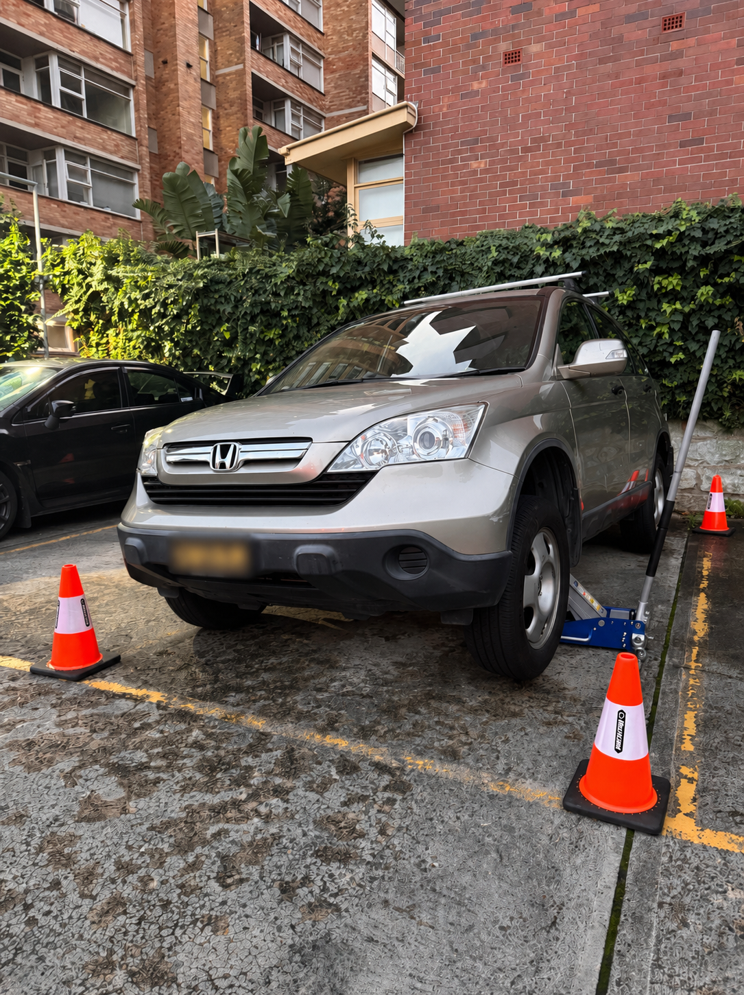 A silver Honda SUV parked in a parking lot with orange traffic cones around it. The front left tire is on a hydraulic lift, indicating the vehicle is being serviced or repaired.