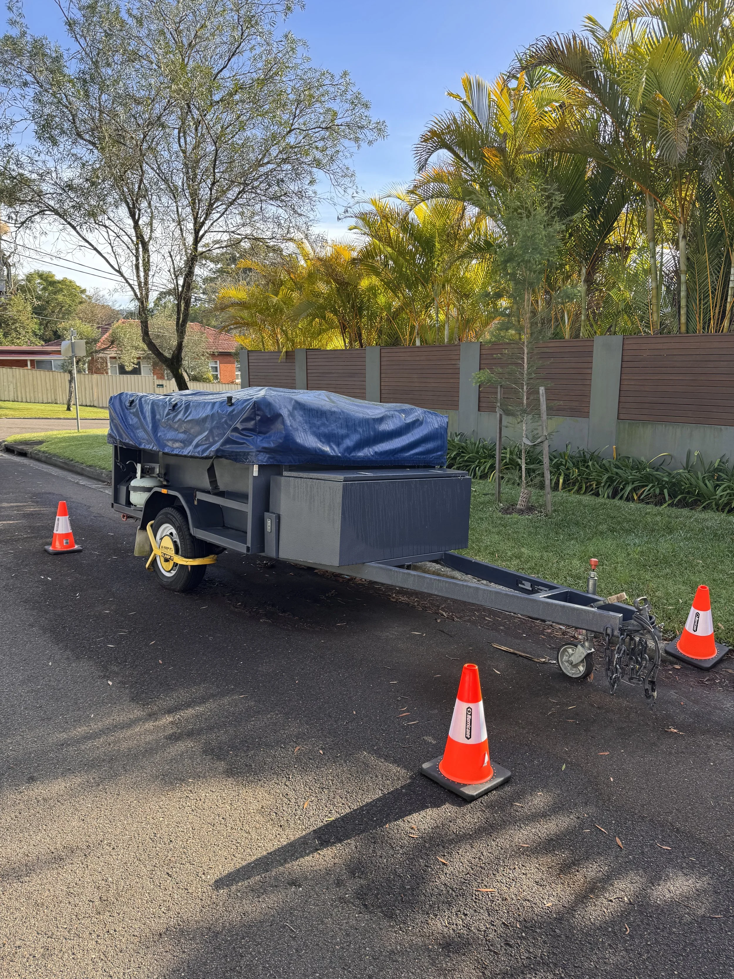 A trailer with a blue cover parked on a freshly paved road, surrounded by orange traffic cones, with trees and a wooden fence in the background.