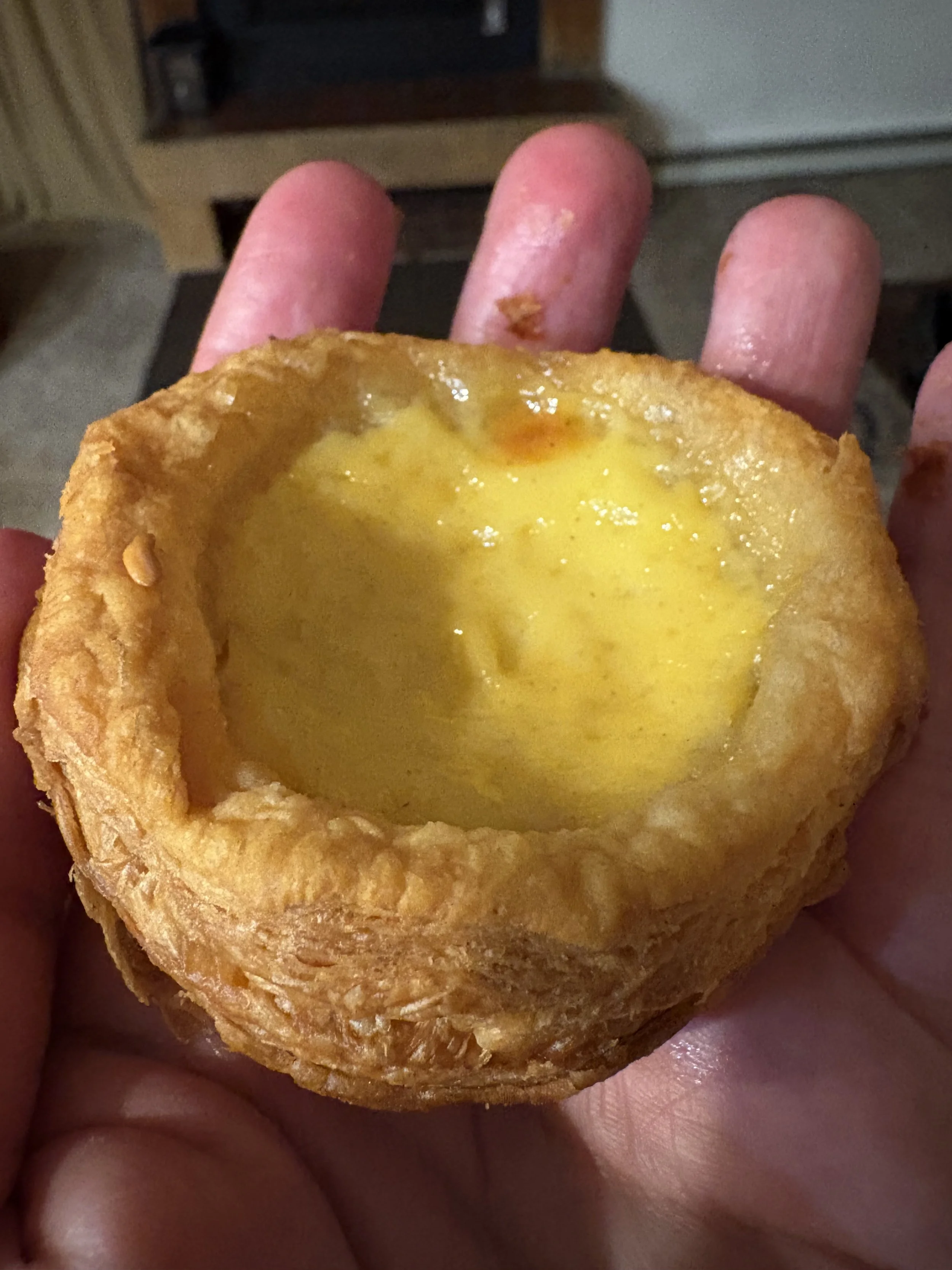 A hand holding an egg tart or pastel de nata with a slightly melted crust and visible bits of food or seasoning.