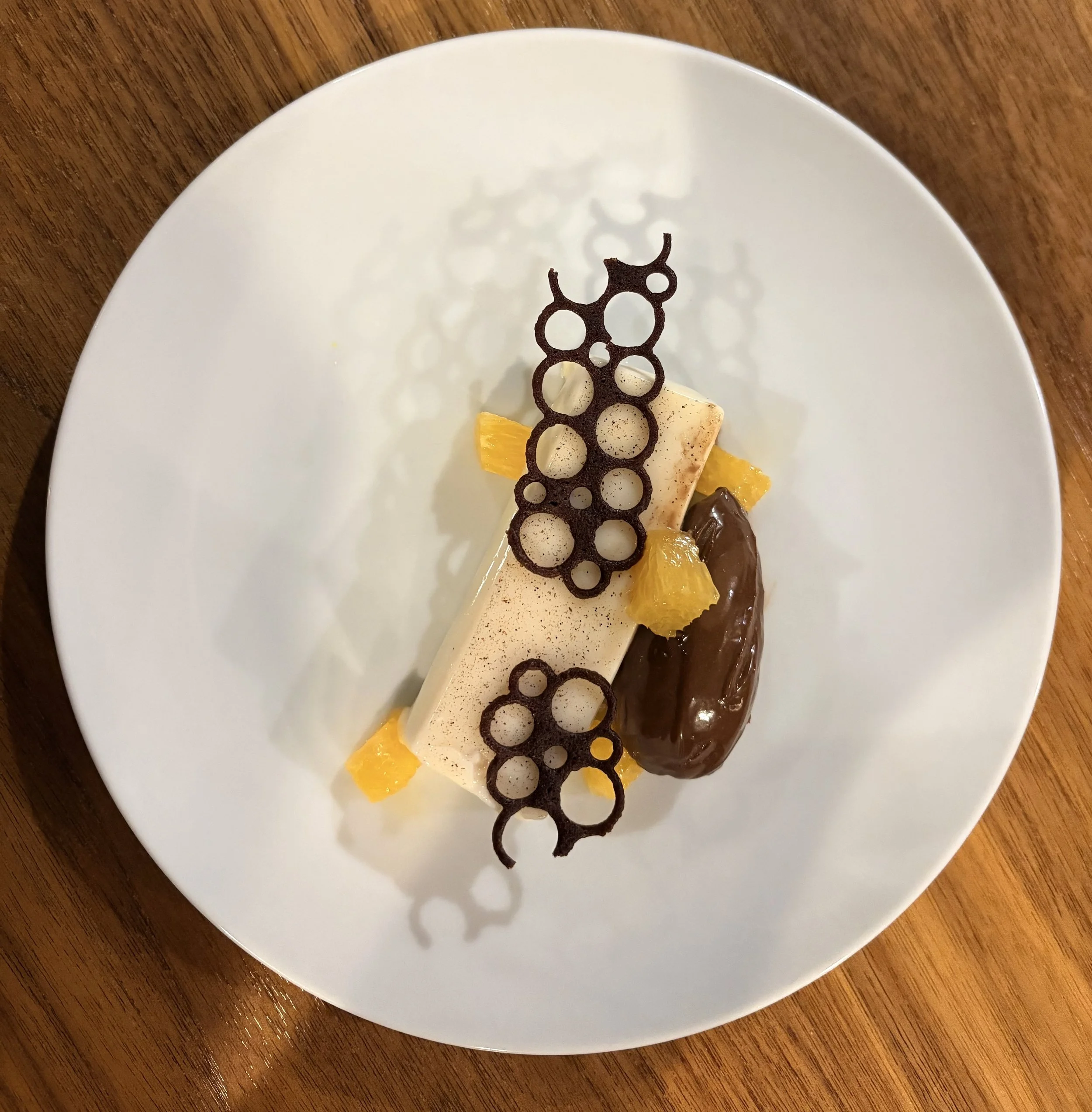Elegant dessert with white mousse, dark chocolate decoration, yellow fruit pieces, and a chocolate glazed eclair on a white plate.