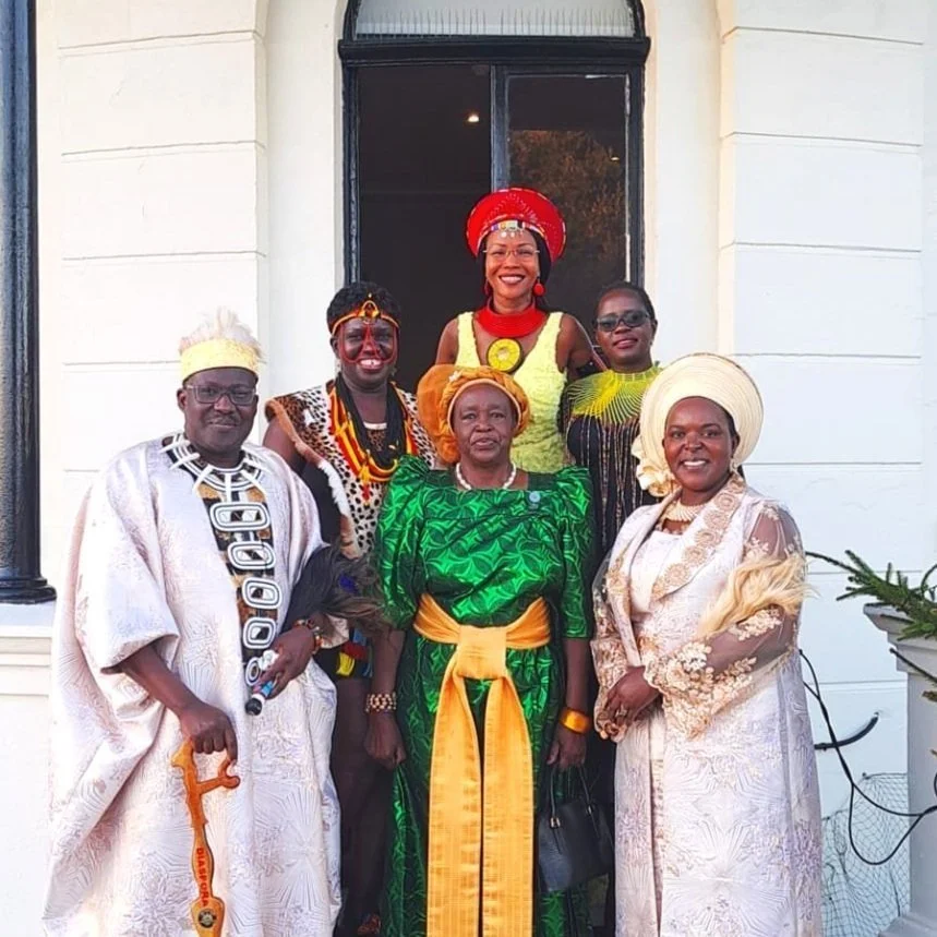 Group of six people dressed in traditional and ceremonial African attire, posing in front of a building with a white exterior, some of them smiling.