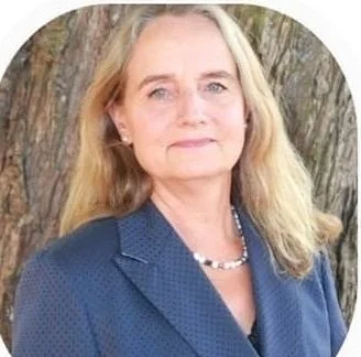 A woman with blonde hair wearing a blue blazer and a necklace, standing outdoors in front of a tree.