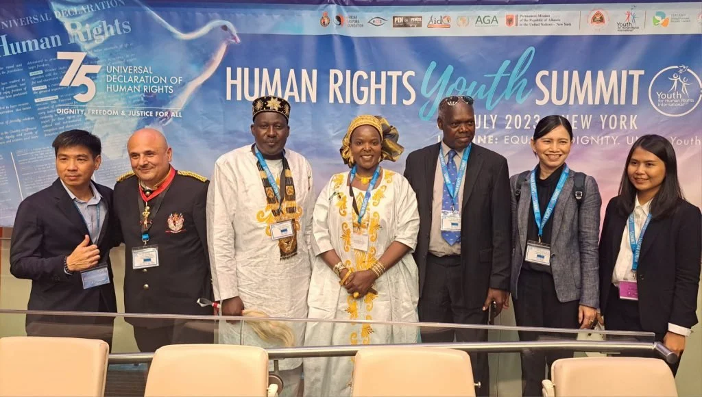 Group of seven diverse people attending the Human Rights Youth Summit, posing for a photo in front of a blue event banner that reads '75 Universal Declaration of Human Rights, Human Rights Youth Summit, July 2023, New York.' They are dressed in formal and traditional attire, all wearing event lanyards.