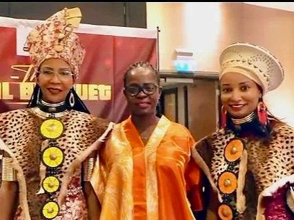 Three women dressed in traditional African attire, standing together at an indoor event.