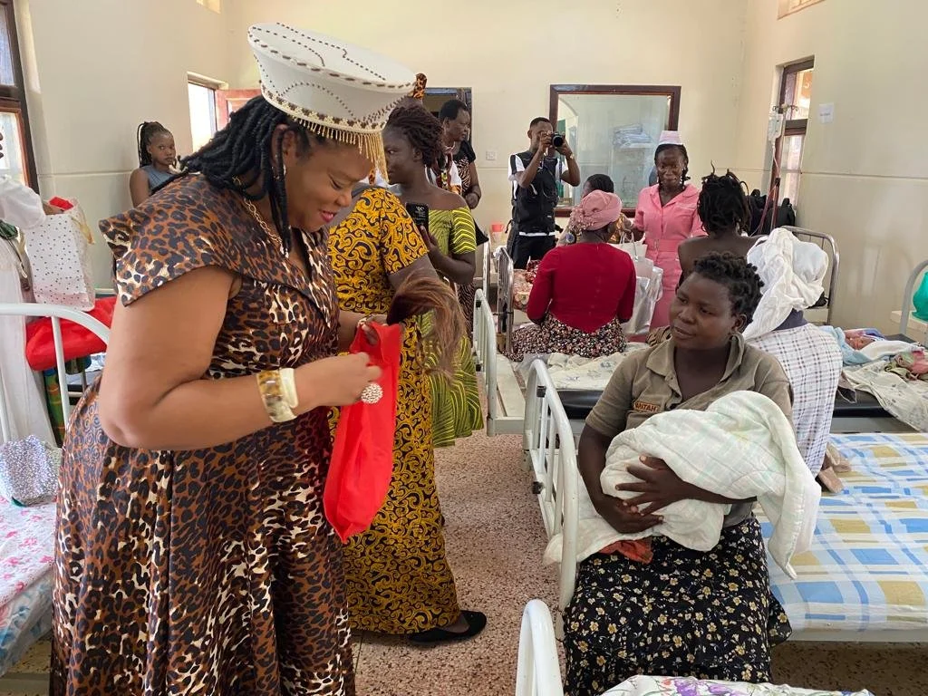 A woman dressed in animal print and wearing a white hat is handing a red bag to a woman sitting on a hospital bed holding a baby wrapped in a blanket, with multiple hospital beds and other women and children in the background.