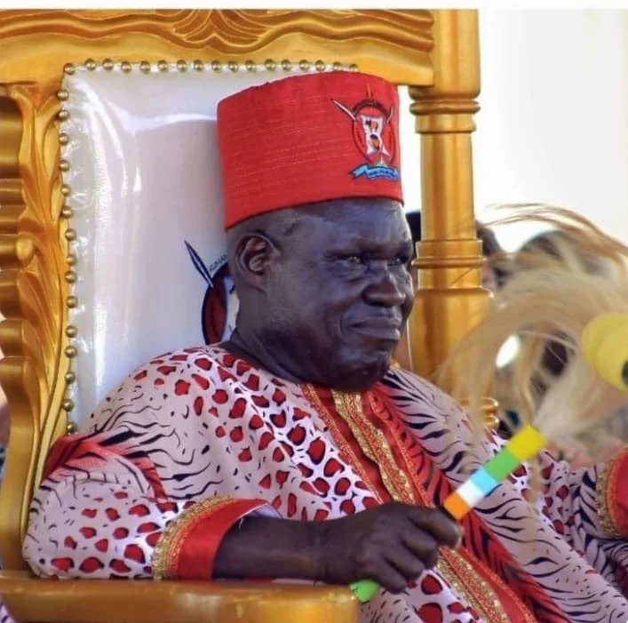 A man dressed in vibrant traditional attire, including a red hat and decorated robe, sitting on an ornate wooden throne.