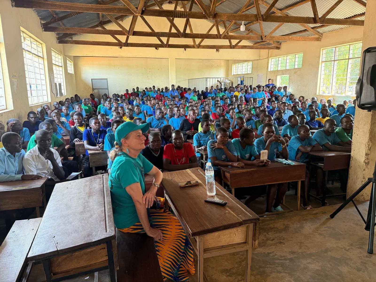 A large classroom filled with students, mostly wearing blue uniforms, attentively listening to a woman sitting at a desk near the front. The room has high windows, wooden ceiling beams, and a simple, rustic interior.