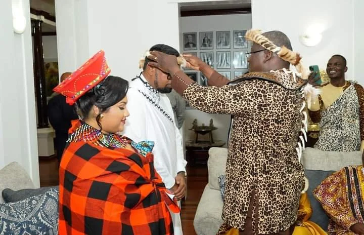People dressed in traditional Maasai attire participating in a ceremony or celebration in a cozy indoor setting, with framed photos on the wall in the background.