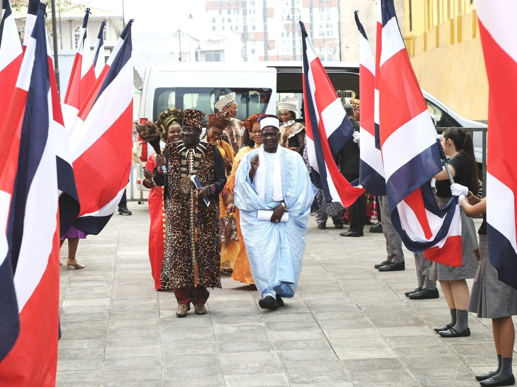Group of people dressed in traditional African attire walking through a corridor of Union Jack flags held by individuals in school uniforms, possibly during a cultural or diplomatic event.