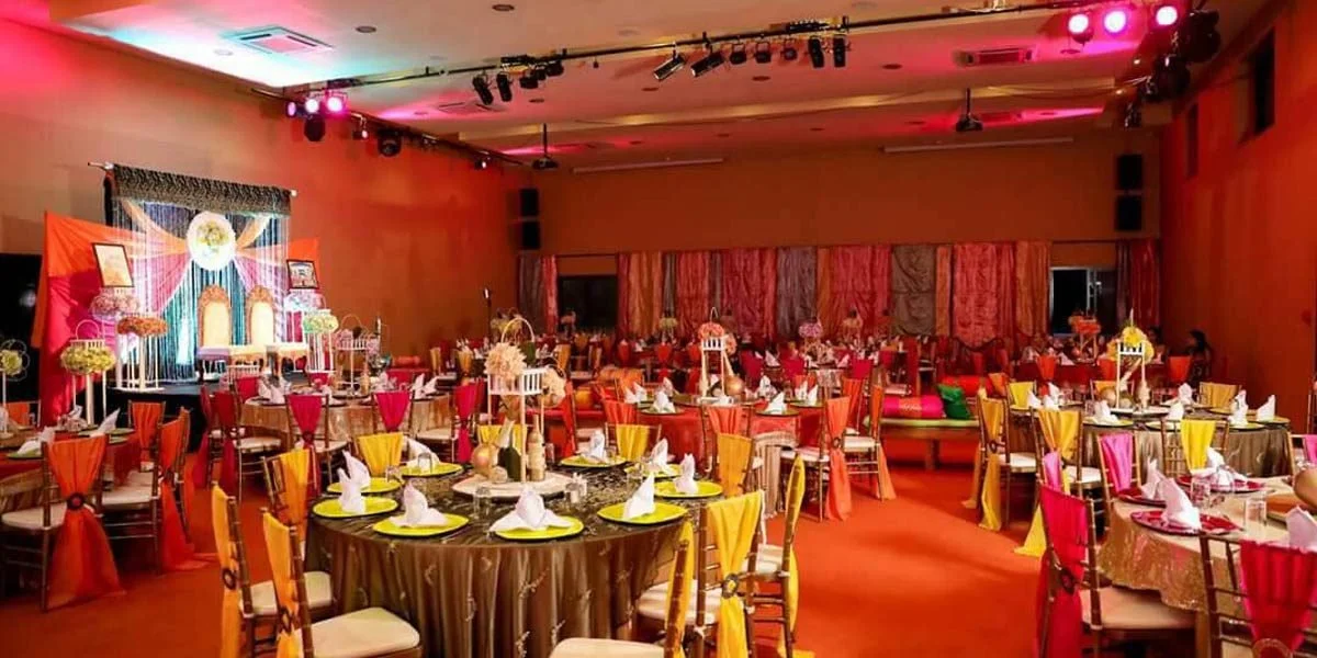 A decorated banquet hall with round tables set for a celebration, adorned with colorful napkins, floral centerpieces, and draped fabric backdrops.