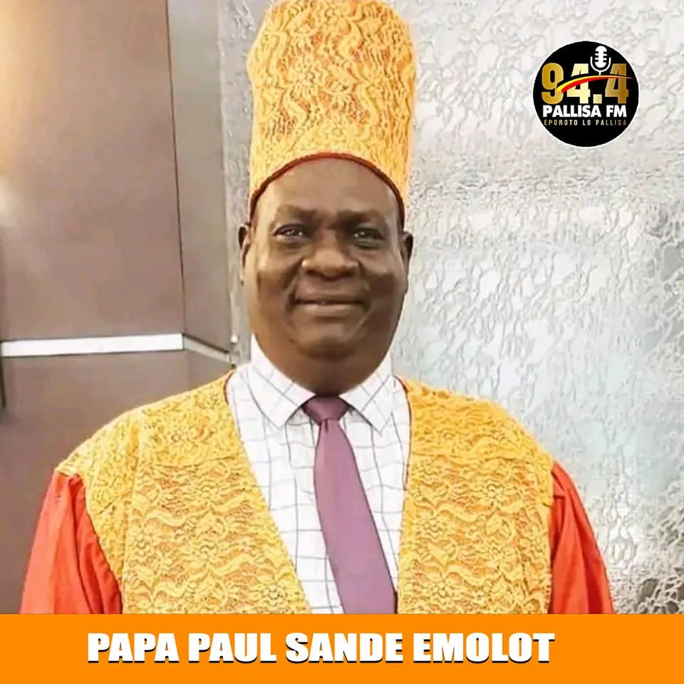 A man dressed in traditional religious attire with a tall, ornate headpiece, smiling for the camera. The image contains a logo for Palusa FM radio station in the top right corner and a caption at the bottom that reads 'Papa Paul Sande Emolot'.